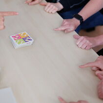 In der Mitte des Tischs liegt eine Spielkarte. Auf ihr sind vier bunte Quadrate abgebildet, in denen jeweils zwei Handformen gezeigt werden. Fünf Spieler machen die Handformen auf der Karte nach.