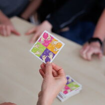 Auf dem Bild ist eine Spielkarte zu sehen, die von einer Hand hochgehoben wird. Auf ihr sind vier bunte Quadrate, in denen jeweils zwei Handformen abgebildet sind. Die Handformen sind in jedem Quadrat gleich.